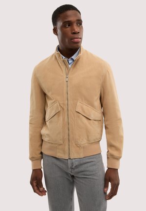Mann trägt eine beige Reißverschlussjacke über einem blauen Hemd mit Kragen und grauen Jeans, steht vor einem schlichten hellen Hintergrund und schaut nach rechts.