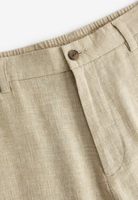 Pantalon en tissu beige texturé avec fermeture à bouton, passants de ceinture et taille partiellement élastique en haut.