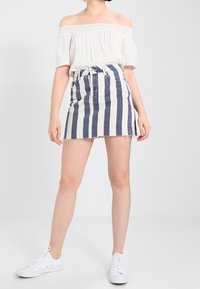 Top blanco de hombros caídos combinado con una mini falda de mezclilla que presenta rayas verticales en azul marino y blanco, con dobladillo deshilachado y cintura ceñida. Zapatillas blancas.