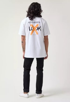 Camiseta de algodón blanca con texto gráfico en negro y naranja en la parte de atrás. Lleva pantalones negros y zapatillas blancas, de pie frente a un fondo neutral.