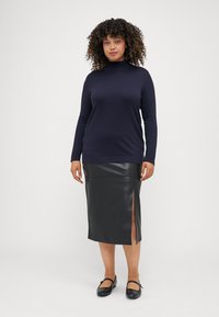 Marineblaues Rollkragenoberteil mit langen Ärmeln; schwarzer Rock aus Kunstleder mit seitlichem Schlitz; schwarze flache Schuhe. Glatte Texturen und kontrastierende Farben.