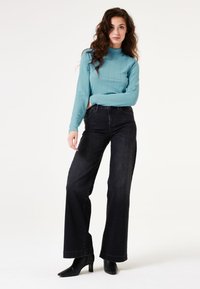 Blau strukturierter Langarmoberteil kombiniert mit hoch taillierten schwarzen Weitbeinjeans und schwarzen Stiefeletten. Vor einem schlichten weißen Hintergrund.