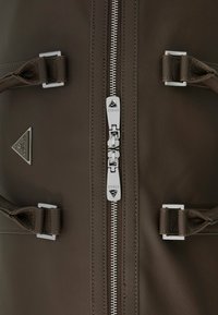 Braune synthetische Materialtasche mit einem zentralen silbernen Reißverschluss, zwei silbernen Schnallen und einem dreieckigen Logo mit "GUESS"-Markierung.