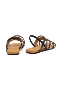 Schwarze Riemchensandalen mit flacher Sohle, aus glattem Material. Mit mehreren überkreuzten Riemen und einer braunen Fußbett. Minimalistisches Design.