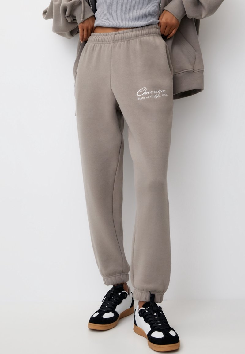 Grå sweatpants med elastisk midja, med "Chicago State of Illinois, USA" tryckt i vitt. Matchas med svarta och vita sneakers.