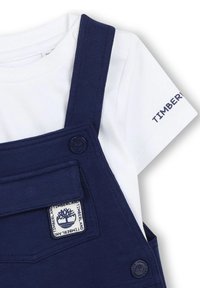 Salopette blu navy con bretelle regolabili, una tasca frontale e una maglietta bianca a maniche corte con il logo "Timberland" sulla manica.