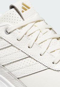 White athletic shoe featuring a textured synthetic upper with perforations, white laces, and three distinct white stripes with gold accents.