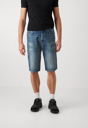 Hellblaue Jeansshorts mit lockerem Schnitt, ausgestattet mit Gürtelschlaufen und Seitentaschen, kombiniert mit einem schwarzen T-Shirt und schwarzen Turnschuhen.