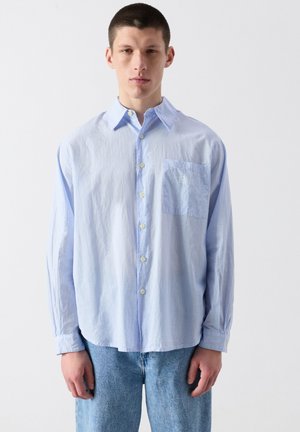 Young man wearing a loose light blue button-up shirt with thin vertical stripes and light blue jeans, standing against a plain background.