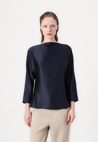 Mørkeblå satengbluse med båthals, trekvartlange ermer og avslappet passform, kombinert med beige bukser. Glatt tekstur og minimalistisk design.