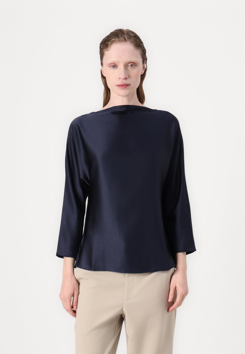 Mørkeblå satengbluse med båthals, trekvartlange ermer og avslappet passform, kombinert med beige bukser. Glatt tekstur og minimalistisk design.