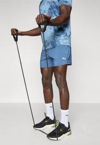 T-shirt de sport à motifs bleus, shorts bleus, chaussettes blanches et baskets noires avec des accents verts. L'utilisateur tient des bandes de résistance de fitness.