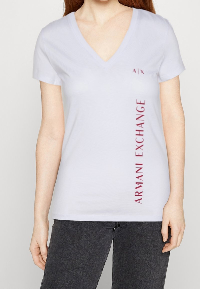 Camiseta blanca de cuello en V hecha de algodón, con el logo "A|X" y el texto "ARMANI EXCHANGE" en burdeos a lo largo del costado. Mangas cortas, tejido ligero.