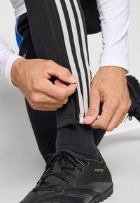 Juodi sportiniai kelnės su baltomis šoninėmis juostelėmis, su užtrauktuko detalumu ties kulkšnimi. Rankos laiko užtrauktuką, derindamos su juodais batais.