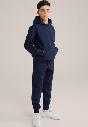 Haut à capuche bleu marine avec une poche kangourou, joggeurs assortis avec des poignets élastiques, portés avec des baskets blanches. Matériau doux en mélange de coton.