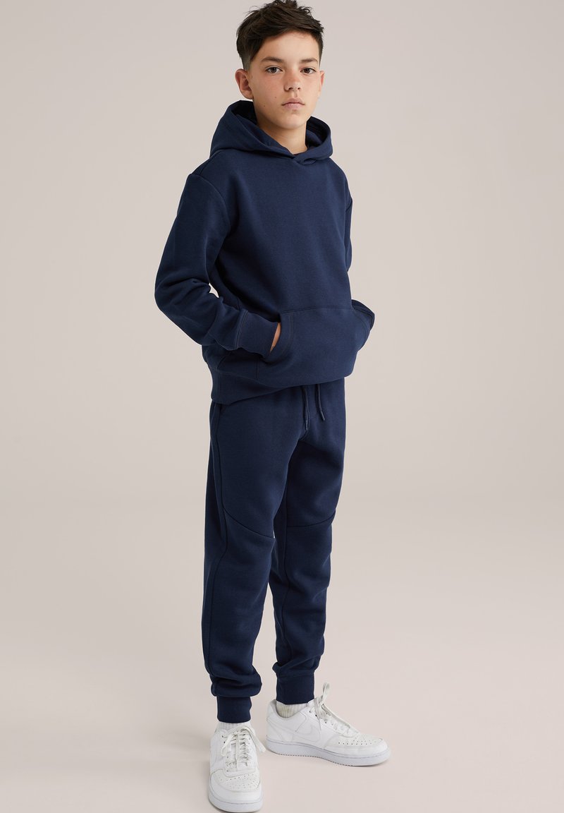 Tumši zili džemperi ar kangaroo kabatu, saskaņoti bikses ar elastīgām piedurknēm, valkātas ar baltām kedām. Mīksts kokvilnas sajaukuma materiāls.