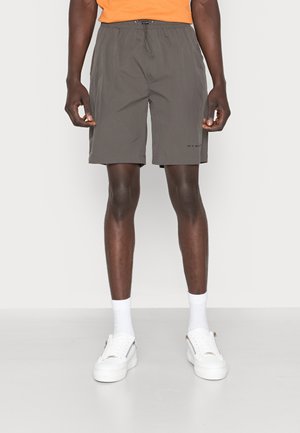 Homme portant un short gris à cordon, des chaussettes blanches montantes et des baskets blanches, debout devant un fond uni.