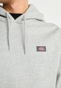 Felpa grigia realizzata in un morbido tessuto, con tasca a marsupio e coulisse regolabile. Presenta una patch con logo rettangolare sul petto.