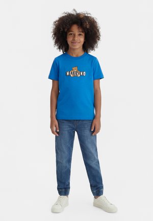 Bambino con capelli ricci che indossa una maglietta blu con un orsetto e il logo "Moschino", jeans blu e scarpe da ginnastica bianche, in piedi e sorridente.
