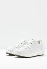 Para białych skórzanych trampek z sznurowaniem, z perforowanymi detalami i grubą, wyściełaną podeszwą na białej powierzchni.