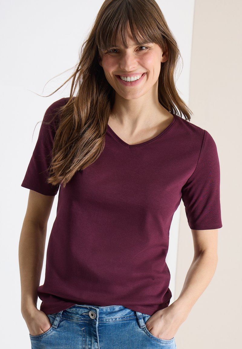 Femme souriante aux longs cheveux bruns portant un t-shirt bordeaux à manches courtes et col en V, ainsi qu'un jean bleu, mains dans les poches.