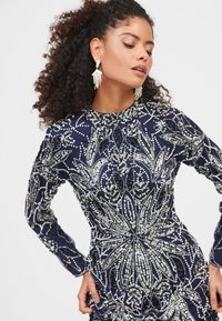 Námořnické šaty s plnými rukávy, zdobené složitými stříbrnými a tmavě modrými korálky ve květinových vzorech; texturovaná látka s vysokým výstřihem.