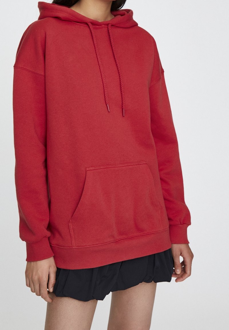 Femme portant un sweat à capuche rouge oversize avec poche avant et une jupe plissée noire, debout devant un fond blanc uni.