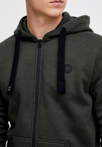 Felpa con zip di colore verde oliva con zip nera, cordini testurizzati e un piccolo logo rotondo sul petto. Tessuto morbido, tasca a marsupio.