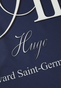 Marineblå stof med hvidt trykt tekst. Der er en elegant serif skrifttype til "Hugo" og en fed sans-serif til "Edward Saint-Germain."