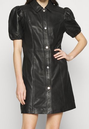 Robe courte en cuir noir avec manches bouffantes et fermeture à boutons-pression à l'avant, portée par une femme avec une main sur la hanche.