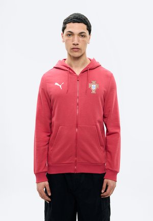 Ung mann med kort krøllet hår iført en rød hettejakke med glidelås, merket med Puma-logo og emblem, stående foran en ensfarget lys bakgrunn.