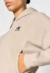 Φούτερ με κουκούλα σε ανοιχτό μπεζ χρώμα από μαλακό ύφασμα, με εμπρόσθιο φερμουάρ, πλαϊνές τσέπες και μικρό μαύρο λογότυπο Champion στο στήθος.