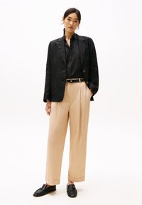 Blazer di lino nero, camicia abbottonata, pantaloni beige a vita alta, mocassini in pelle nera. Design minimalista con forme rettilinee e trame sottili.