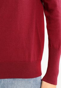 Gros plan sur une personne portant un pull à manches longues rouge avec des poignets et un ourlet côtelés, associé à un jean en denim bleu clair.