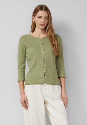 Frau mit langen blonden Haaren, die eine hellgrüne, geknöpfte Strickjacke und cremefarbene Hose trägt, steht vor einem schlichten grauen Hintergrund.