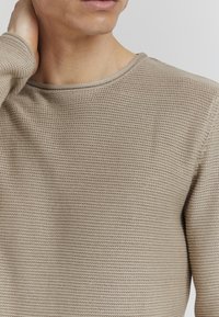Beige Langarmstrickshirt mit strukturiertem Oberflächen und rundem Ausschnitt, das dezente Nähte entlang des Ausschnitts und der Ärmel aufweist.