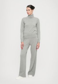Grau gestrickter Rollkragenpullover und weit geschnittene Hose, beide mit Rippdetails. Das Modell trägt weiße Sneakers und steht vor einem schlichten Hintergrund.