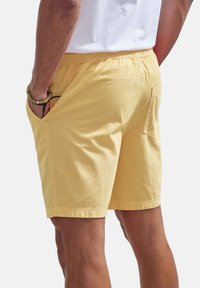 Yellow cotton shorts with an elastic waistband, side pockets, and a back pocket. Smooth texture and slightly tailored design.