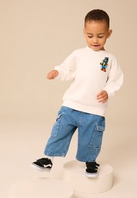 Hanorac alb cu grafică colorată, pantaloni scurți cargo albastru deschis și adidași negri cu talpă albă. Copilul stă pe piedestale albe.