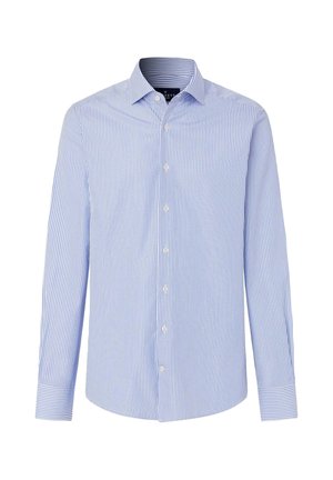 Hombre de pie con una camisa de rayas azul claro abotonada, cinturón negro, pantalones negros y zapatos de cuero negro sobre un fondo claro y liso.