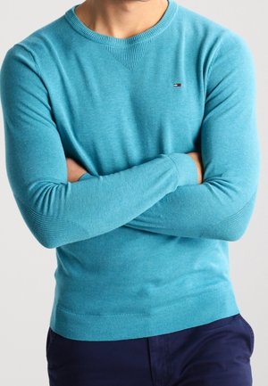Uomo che indossa un maglione aderente turchese a scollo a V con piccolo logo, braccia incrociate, abbinato a pantaloni blu scuro su sfondo neutro.