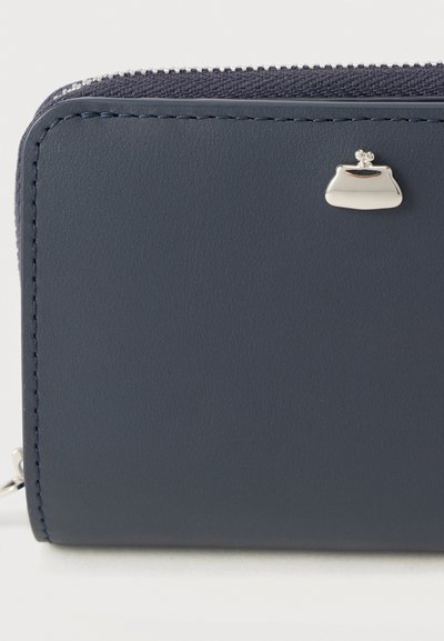 Πορτοφόλι από σκούρο μπλε δέρμα με ομαλή υφή και ασημένια αγκράφα. Κλείνει με φερμουάρ στην κορυφή και έχει αντίθετη ραφή στα άκρα.