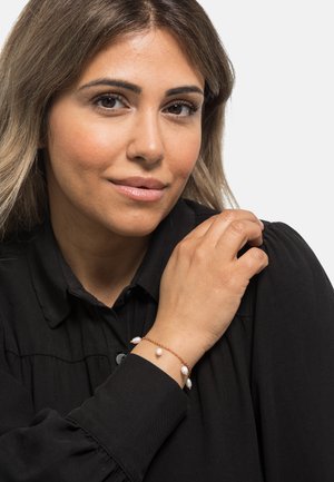 Bracciale con catena in oro rosa e molteplici accentuazioni di perle bianche. La modella indossa una blusa nera con colletto, mettendo in risalto i gioielli.