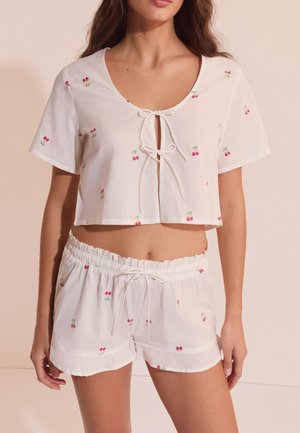 Une femme porte un ensemble de pyjama deux pièces blanc avec de petits motifs de cerises rouges, manches courtes, haut court avec liens à l'avant, et short taille élastique.