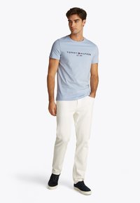 Light blue cotton t-shirt with a crew neckline and printed logo in black, paired with white pants and navy suede shoes.