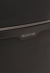 Sac en cuir noir avec une finition texturée, doté d'une fermeture éclair argentée et d'une plaque logo. Forme rectangulaire compacte.