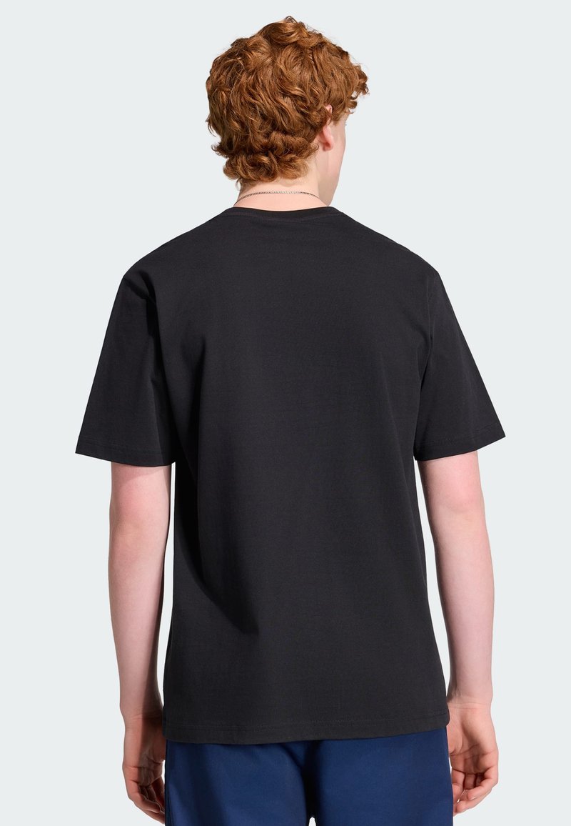 Junge Person mit lockigem roten Haar, die ein schlichtes schwarzes T-Shirt mit kurzen Ärmeln und blaue Hose trägt, mit dem Rücken zum Betrachter vor einfarbigem Hintergrund.