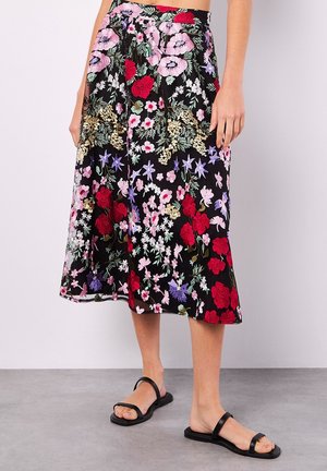 Woman wearing a black midi skirt with red, pink, purple, and white floral embroidery and black flat sandals with two straps.