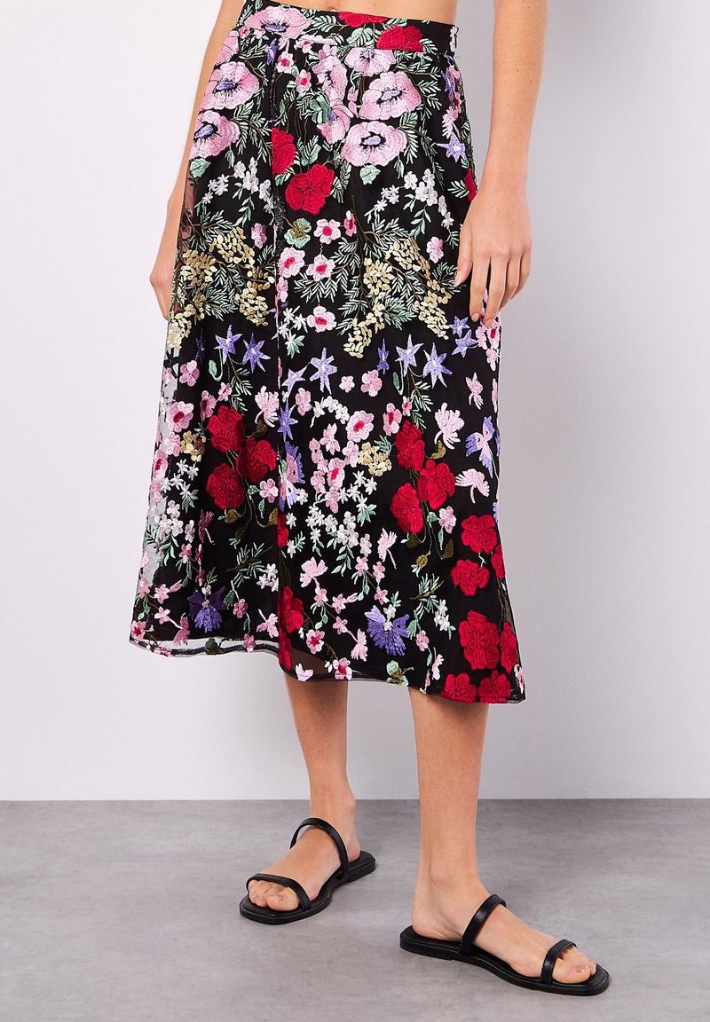 Femme portant une jupe midi noire avec une broderie florale rouge, rose, violette et blanche, et des sandales plates noires à deux brides.