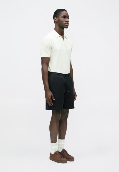 Polo à manches courtes vert clair avec un col zippé, associé à un short noir, des chaussettes blanches montantes et des chaussures marron. Design minimaliste.